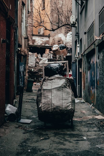 A narrow urban alleyway filled with a significant amount of dumped waste and refuse. In the foreground, a large, grey, weathered bag or sack, possibly made of fabric or heavy-duty plastic, is positioned on the uneven, dark asphalt surface. The sack appears to be partially filled, with visible creases and wrinkles. Behind it, numerous cardboard boxes and plastic bags are stacked haphazardly on a makeshift pile, with some boxes torn or crushed. The background features a clutter of various waste materials, including shredded paper, plastic containers, and miscellaneous debris, stretching up a small incline or wall surrounded by leafless tree branches and bare brick or concrete structures. The alley is flanked by narrow buildings, with graffiti on the walls to the right and boarded-up or darkened doorways on the left. The scene is lit by natural daylight, casting subtle shadows and highlighting the textures of the rubbish and surrounding surfaces. This image exemplifies the need for private waste clearance services, such as those provided by Rubbish Clearance Hammersmith, for effective on-site rubbish removal and alternative waste handling outside of municipal collection.