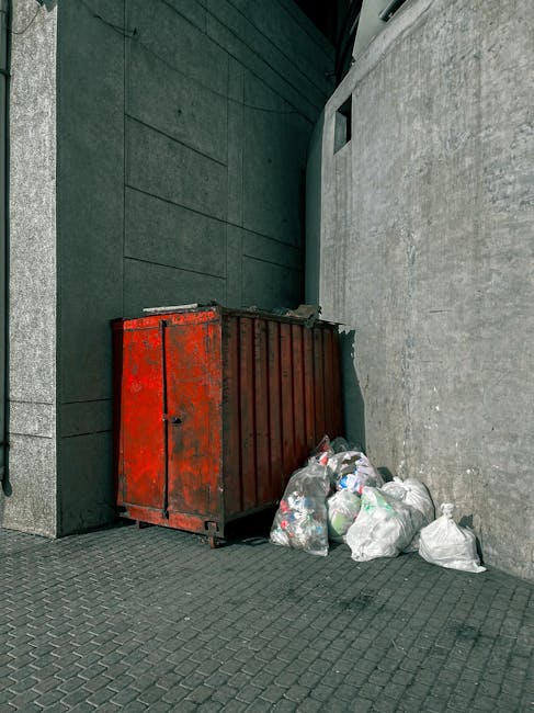 A weathered red metal dumpster with visible rust and peeling paint, positioned against a dark green wall with a textured finish and adjacent to a light grey concrete wall. The dumpster is situated on a paved surface composed of small, interlocking dark grey bricks. Several plastic garbage bags filled with mixed waste are placed next to the dumpster on the ground, some leaning against its side. The environment appears to be an outdoor urban alley or service area, with a small rectangular opening in the green wall and ambient lighting suggesting daytime. The scene illustrates a typical setting for private rubbish collection or waste disposal outside commercial or residential buildings, aligning with the context of independent rubbish removal services provided by companies such as Rubbish Clearance Hammersmith.