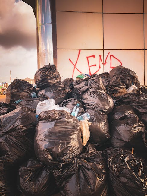 A narrow urban alleyway filled with a significant amount of dumped waste and refuse. In the foreground, a large, grey, weathered bag or sack, possibly made of fabric or heavy-duty plastic, is positioned on the uneven, dark asphalt surface. The sack appears to be partially filled, with visible creases and wrinkles. Behind it, numerous cardboard boxes and plastic bags are stacked haphazardly on a makeshift pile, with some boxes torn or crushed. The background features a clutter of various waste materials, including shredded paper, plastic containers, and miscellaneous debris, stretching up a small incline or wall surrounded by leafless tree branches and bare brick or concrete structures. The alley is flanked by narrow buildings, with graffiti on the walls to the right and boarded-up or darkened doorways on the left. The scene is lit by natural daylight, casting subtle shadows and highlighting the textures of the rubbish and surrounding surfaces. This image exemplifies the need for private waste clearance services, such as those provided by Rubbish Clearance Hammersmith, for effective on-site rubbish removal and alternative waste handling outside of municipal collection.