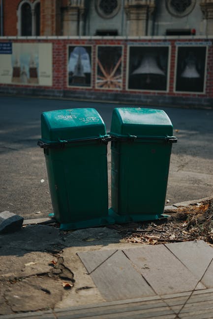 Two green wheelie bins with hinged lids, positioned side by side on the pavement near the edge of a street. The bins are made of durable plastic with a matte finish, displaying some minor signs of weathering. They are placed directly on an uneven surface that includes concrete paving slabs and a small area of dirt and dried leaves around their base. Behind the bins, there is a road with asphalt surface, which appears to be lightly used, and across the street, there is a building with a brick and stone exterior, featuring windows with decorative ironwork and arch details. In the background, a segment of sidewalk and a construction barrier with posters or advertisements are faintly visible. The scene is illuminated by natural daylight, contributing to clear visibility of the surrounding urban environment. This setup reflects an alternative waste management approach where individual disposal units are positioned for collection, aligning with private rubbish removal services provided by companies like Rubbish Clearance Hammersmith, especially in areas where on-site clearance or independent rubbish disposal is utilized.