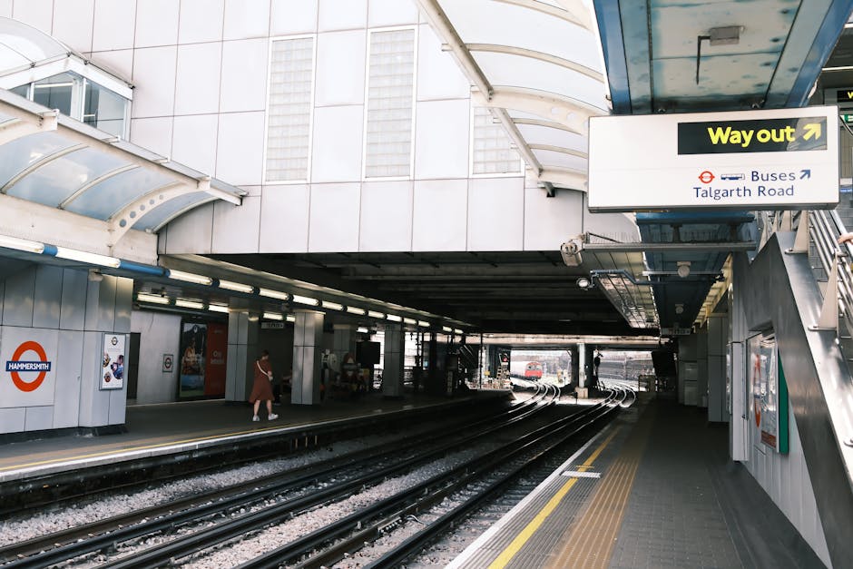 The image depicts a modern underground train station platform with a curved, translucent canopy overhead. On the left side, there are white wall panels with the London Underground roundel logo and some posters. The platform surface appears concrete with tactile paving near the edge for safety. A woman dressed in casual clothing is walking toward the escalator or stairs on the left, while other passengers are seated further along the platform, under a row of linear lighting fixtures. The railway tracks run centrally, curving into the station’s distance, with a train visible further back on the right side. The environment is well-lit with natural light filtering through the canopy and artificial lighting, creating a neutral, functional atmosphere typical of urban transit stations. Although primarily focusing on public transport infrastructure, the setting subtly underscores the importance of efficient waste disposal and management in busy areas, relevant to services like those provided by Rubbish Clearance Hammersmith, which may facilitate alternative waste handling solutions outside conventional public disposal channels in similar environments.