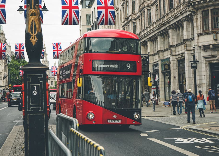 A red double-decker bus is driving along a busy London street, with the route number 9 and destination Hammersmith displayed on the front. The bus is positioned in the center of the image, with its sleek, modern design and large tinted windows. On the left side, a black lamppost adorned with Union Jack flags is visible, adding a patriotic decorative element, and a metal barrier runs parallel to the curb. In the background, there are historic stone buildings with ornate architectural details, large windows, and decorative cornices, typical of central London. Pedestrians, some with backpacks and coats, are walking along the sidewalk, with a mix of casual and business attire. The environment appears lively but orderly, with natural daylight illuminating the scene, reflecting typical urban surroundings. Although the primary focus is the bus, this scene relates to urban transportation in London, an alternative to private waste handling and on-site clearance services, which Rubbish Clearance Hammersmith may serve in adjacent areas.
