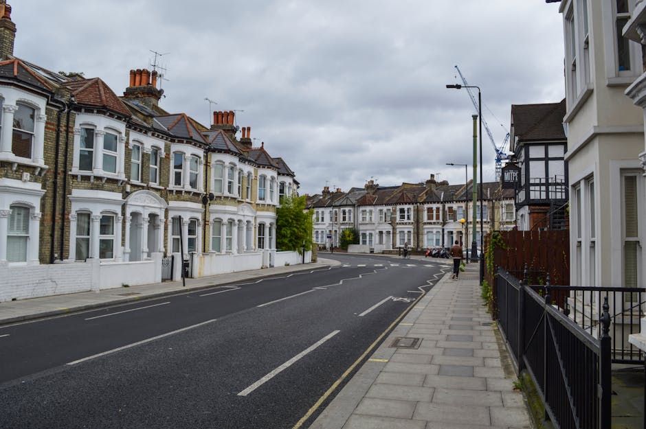 A quiet residential street featuring a row of Victorian-style terraced houses with ornate bay windows, decorative brickwork, and white-painted facades. The street is paved with tarmac and marked with white lines for parking spaces, although no vehicles are presently parked. A narrow, clean concrete sidewalk runs alongside, bordered by black metal railings and a few small plants. Overhead, the sky is cloudy, casting diffuse light over the scene. To the right, a modern building with white walls and black trim stands close to the sidewalk, with a wooden fence partially visible. Trees with fresh green leaves are interspersed along the street, adding a touch of nature to the urban setting. The area appears well-maintained and quiet, typical of an inner-city residential zone where private waste collection services, such as those offered by Rubbish Clearance Hammersmith, might be employed to manage rubbish removal, especially for estate or property renovation projects. The scene captures the orderly, typical appearance of a fashionable London suburb that may require regular rubbish clearance for upkeep or refurbishment.