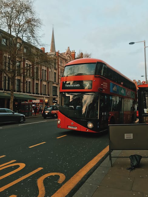 A double-decker bus in red and black livery, displaying the route number 9 and destination Hammersmith, is positioned on a city street with a clear view of the front and partial side of the bus. The bus is situated on the right side of the image, adjacent to a paved sidewalk that features a bench and a black bag resting on the ground. Behind the bus, several cars are seen moving along the roadway, which is marked with yellow lines. The background reveals a row of multi-storey brick buildings with large windows, some featuring commercial storefronts at street level. Leafless trees line the sidewalk, indicating a late autumn or winter season, and a tall church spire is visible against a pale, overcast sky. The lighting suggests either early morning or late afternoon, with soft, diffuse natural light creating gentle reflections on the bus's glossy surface. The scene captures an urban environment typical of central Hammersmith, with a subtle connection to the local transportation network and the context of civic or private travel, aligning with the themes of independent public transport or alternative arrangements for city upkeep.