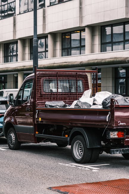 A small, dark red pickup truck is parked on a city street in front of a modern, multi-story building with large rectangular windows and a concrete facade. The truck's cargo bed is loaded with various types of rubbish, including black and white plastic bags, cardboard, and other waste materials. The truck has a protective metal grille at the back of the cab, and the side and rear panels are smooth with a slightly glossy finish. Surrounding the vehicle, the street features painted road markings indicating parking spaces, and a tall street lamp is positioned nearby. In the background, other vehicles, including a white van, are visible, along with reflections of the urban environment in the building's windows. This scene depicts a typical private rubbish collection scenario, possibly handled by a waste management service such as Rubbish Clearance Hammersmith, highlighting the process of on-site clearance of waste materials before disposal or recycling.
