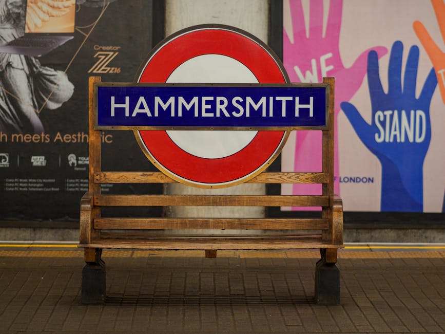 A double-decker bus in red and black livery, displaying the route number 9 and destination Hammersmith, is positioned on a city street with a clear view of the front and partial side of the bus. The bus is situated on the right side of the image, adjacent to a paved sidewalk that features a bench and a black bag resting on the ground. Behind the bus, several cars are seen moving along the roadway, which is marked with yellow lines. The background reveals a row of multi-storey brick buildings with large windows, some featuring commercial storefronts at street level. Leafless trees line the sidewalk, indicating a late autumn or winter season, and a tall church spire is visible against a pale, overcast sky. The lighting suggests either early morning or late afternoon, with soft, diffuse natural light creating gentle reflections on the bus's glossy surface. The scene captures an urban environment typical of central Hammersmith, with a subtle connection to the local transportation network and the context of civic or private travel, aligning with the themes of independent public transport or alternative arrangements for city upkeep.