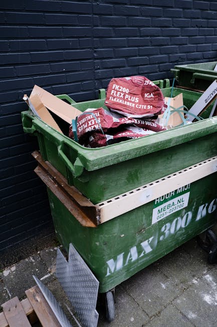 A large green wheelie bin filled with several heavy-duty black and red bags of construction or landscaping waste, identified by visible text on the bags, stacked on top of another green bin placed on a paved surface near a dark blue brick wall. The bins are positioned on a concrete pavement with some debris and a metal grate nearby, indicating an outdoor area potentially used for waste collection or disposal. The scene appears to be in an urban or commercial setting, with the presence of multiple bins and collected materials suggesting an activity related to rubbish removal or private waste management services, as provided by Rubbish Clearance Hammersmith. The environment is lit by natural daylight, with shadows cast on the ground and a clean but utilitarian atmosphere typical of a site preparing for waste collection or disposal.