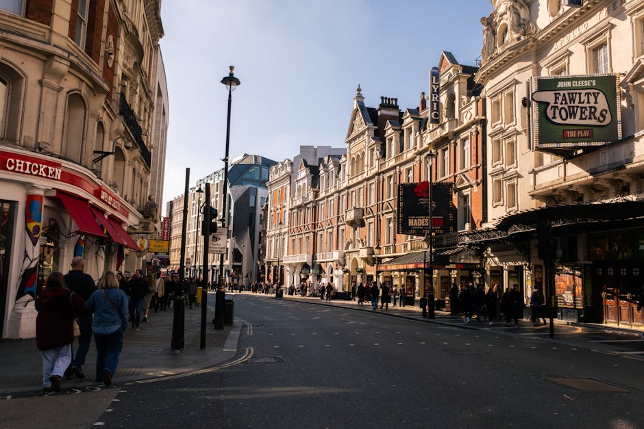 A busy city street scene featuring a mixture of historic and modern buildings with ornate facades, tall windows, and decorative detailing, lining both sides of the road. On the left, there is a storefront with a red and white awning displaying the word 'CHICKEN,' attracting pedestrians walking along the sidewalk. Several people are visible, some walking in groups and others alone, dressed in casual clothing, and heading towards various shop entrances or crossing the street. Black bollards and street lamps are positioned along the pavement, which appears clean and well-maintained. On the right side, buildings have a variety of signage, including a large illuminated sign for 'Fawty Towers' theatrical venue, and smaller signs for other businesses, suggesting a vibrant commercial area. The road itself is relatively clear, with lane markings visible, and no vehicles in sight. The lighting indicates a daytime setting with a partly cloudy sky, creating a natural and neutral atmosphere suitable for describing urban rubbish removal and waste management services in a lively city environment.