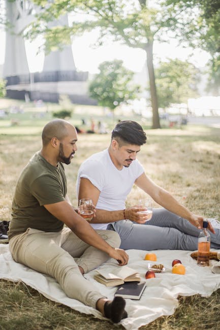 Two men sitting on a white blanket spread on the grass in a park, with trees and a modern building in the background, enjoying drinks and snacks. One man, with a shaved head and beard, is wearing a green t-shirt and beige trousers, holding a glass of what appears to be a beverage. The other man, with dark hair and a clean-shaven face, is dressed in a white t-shirt and grey trousers, reaching for a bottle of a drink. Between them, there are apples and a book on the blanket, creating a relaxed picnic scene. The scene is outdoors with natural light filtering through the leafy trees, and the setting suggests a peaceful, recreational environment. This image connects subtly to the theme of waste management by illustrating a clean and organized outdoor setting, akin to the controlled disposal or clearance activities handled by services like Rubbish Clearance Hammersmith, which provides alternative waste handling options outside of local authority collection.