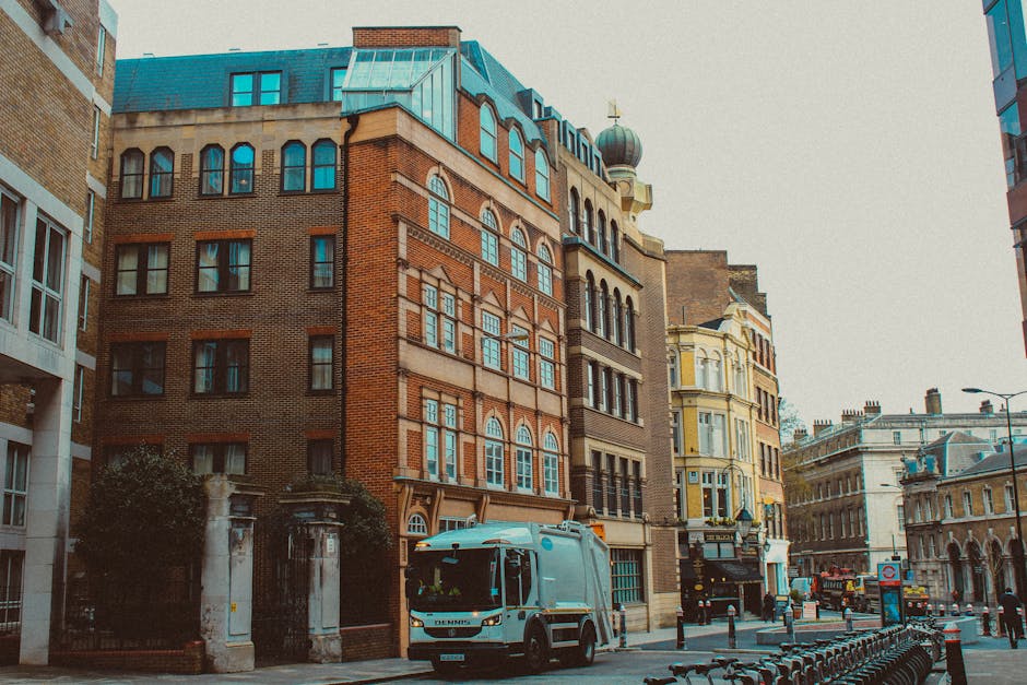 A street scene in an urban area showing a mix of architectural styles in multi-storey buildings, including a prominent red-brick structure with arched windows and decorative stonework, alongside other buildings with yellow and beige facades. A light blue rubbish collection truck is parked on the street in the foreground, with visible details such as the cab and compaction area. The street is lined with metal bollards and bike racks, and a sidewalk with some greenery and trees is partially visible on the left. In the background, additional historic and modern buildings form a busy cityscape, with traffic signs and streetlights present, indicating an active thoroughfare. The scene is set during daylight with overcast lighting, contributing to a neutral and professional atmosphere consistent with urban rubbish removal and waste management efforts, such as those undertaken by Rubbish Clearance Hammersmith.